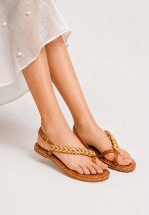 Voeten met bruine platte sandalen met gevlochten bandje, gecombineerd met een witte rok versierd met kleine parels.