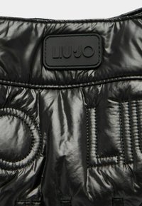 Fekete, steppelt nejlon dzseki részlete, amely texturált anyagot és dombornyomott logócímkét tartalmaz. Simább felület vízszintes és körkörös varrással.