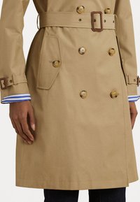 Beige trench coat with double-breasted buttons, belt, and a side pocket; features blue and white striped cuff detail.
