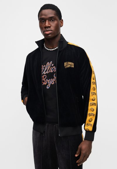 Μαύρο βελούδινο μπουφάν με φερμουάρ και κίτρινες πλευρικές ρίγες, με λογότυπα "Billionaire Boys Club", συνδυασμένο με μαύρο graphic T-shirt.