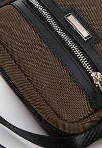 Borsa a tracolla in tessuto marrone con texture, dettagli in pelle nera, zip argentata e etichetta con logo. Presenta una tracolla staccabile per maggiore versatilità.