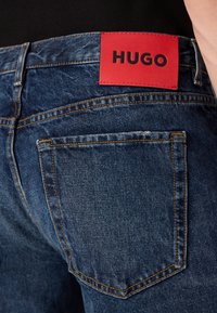Džínsy z denimu s tmavým praním, s oranžovým prešívaním, s obdĺžnikovou červenou koženou nášivkou označenou "HUGO" na páse a s jednou zadnou vreckom.