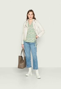 Jaqueta de moto em pele sintética de cor clara sobre uma blusa floral verde, combinada com jeans de perna reta de tom azul claro e botas brancas de salto. Bolsa tote castanha.