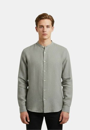 Uomo che indossa una camicia grigio chiaro a maniche lunghe con collo alla coreana, in piedi davanti a uno sfondo bianco semplice.