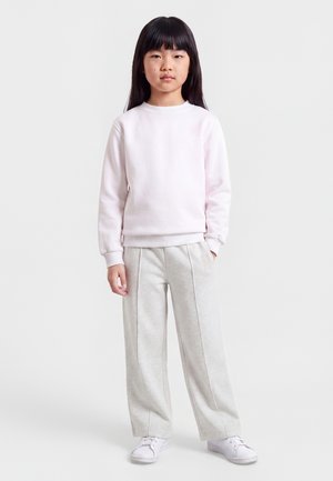 Jeune fille aux longs cheveux noirs portant un sweat-shirt rose pâle, un pantalon large gris clair et des baskets blanches, debout avec une main dans la poche.