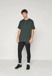 Nuorodinis vyras stovi vilkėdamas tamsiai žalią per didelį marškinėlį, juodus Nike kelnes, baltes kojines ir juodus sportbačius prieš paprastą šviesią foną.