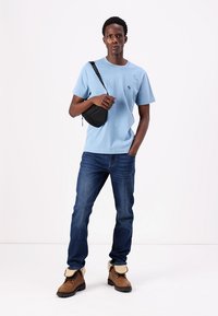 Light blue cotton t-shirt with a small logo, worn with blue denim jeans and brown boots featuring a beige collar. Black crossbody bag.
