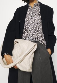 Borsa a tracolla intrecciata di colore beige con una finitura morbida e testurizzata, tenuta sotto un cappotto oversize nero, abbinata a una blusa leggera a fantasia e pantaloni grigi.
