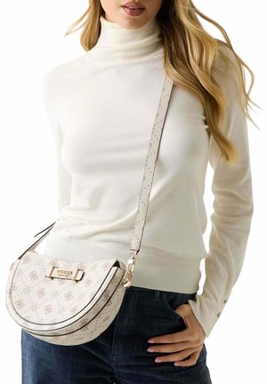 Femme portant un col roulé crème et un jean foncé, portant un sac bandoulière blanc à motif avec le logo doré "GUESS" à l'avant.