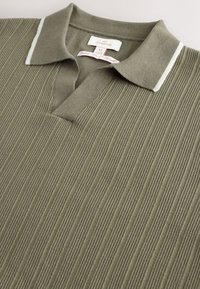 Tricou polo kaki cu textura ridată, guler scurt și dungi albe accentuate pe guler. Prezintă o deschidere la gât cu nasturi.
