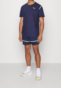 Merkkiväriinen urheilupaita, jossa on raidallinen rakenne, lyhyet hihat ja neonvihreät yksityiskohdat, yhdistettyinä matching-shortsien kanssa ja valkoisten, värikkäiden juoksukenkien kanssa.