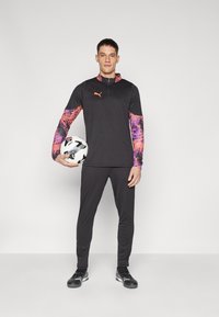 Puma athletic outfit in black with bright pink and purple patterned sleeves. Model holds a soccer ball and wears soccer shoes.