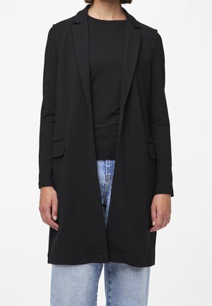 Personne portant un long blazer noir ouvert sur une chemise noire et un jean bleu clair, debout devant un fond blanc uni.