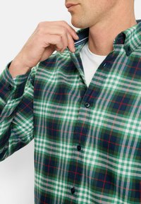 Grünes, marineblaues und rotes Karomuster-Hemd aus Flanellstoff. Verfügt über einen Button-Down-Kragen und Frontknöpfe, mit einem sichtbaren weißen Unterhemd.