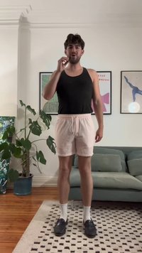 Top in cotone nero, pantaloni corti a scacchi rosa chiaro con cordoncino, calze bianche e mocassini neri. Ambiente interno con pianta verde e divano.