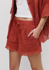 Shorts en crochet couleur rouille avec une taille élastique, un ourlet festonné et une broderie florale. Associés à une chemise à mailles ouvertes assortie.
