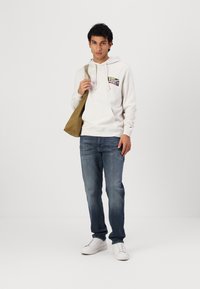 Lys grå hettegenser med et grafisk logo, blå jeans og hvite sneakers. Brune lerret skulderveske kompletterer antrekket.