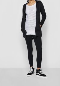 Sweat à capuche noir zippé, haut en tricot texturé blanc, leggings noirs et baskets montantes noires avec accents blancs. Design simple et fonctionnel.
