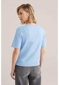 Vaaleansininen teksturoitu lyhythihainen villapaita, jossa on geometrinen kuvio ja resorit hihansuissa ja helmassa, yhdistetty harmaisiin korkeavyötäröisiin farkkuihin.