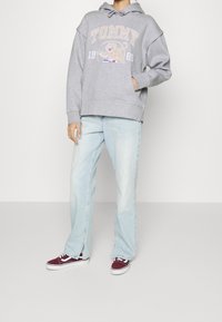Felpa grigia con grafica "TOMMY" e leone, abbinata a jeans azzurri a gamba dritta e sneaker in tela maroon con accenti bianchi.