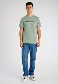 Vihreä puuvillapaita, jossa on laivastonsinisin painatuksin "COPENHAGEN LINDBERGH", yhdistettynä sinisiin farkkuihin ja valkoisiin lenkkareihin. Yksinkertainen, rento muotoilu.