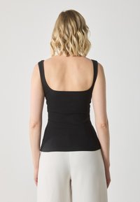 Top sin mangas negro con revestimiento acanalado y escote cuadrado, de corte ajustado, tirantes gruesos y textura suave, combinado con pantalones de color claro.