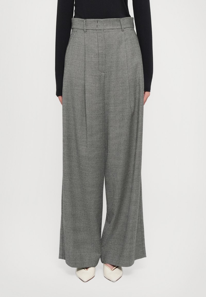 Pantalons larges plissés en motif pied-de-poule gris, dotés d'une taille ajustée, de passants de ceinture et d'une texture lisse. Associés à des talons blancs.