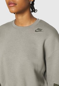 Grå sweatshirt med rund halsringning, mjuk textur och en grön Nike-logotyp på vänster axel. Enkelt mönster med sänkta axlar.