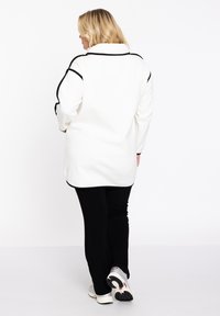 Lange witte fleecjas met zwarte pipingdetails, gecombineerd met zwarte broeken en sportieve schoenen, gezien vanaf de achterkant.
