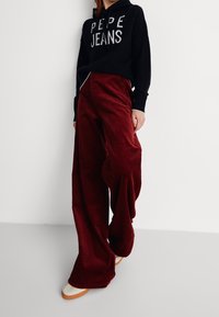 Sweat à capuche noir avec le logo "PEPE JEANS", associé à un pantalon large en velours côtelé bordeaux. Le pantalon présente une texture côtelée verticale et des poches latérales.