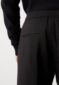 Close-up of person wearing black pants with elastic waistband and back welt pocket, paired with a long-sleeve black top.
