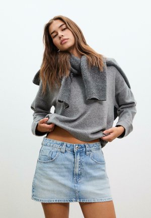 Junge Frau in einem hellblauen Jeans-Minirock und grauem Pullover mit passendem Pullover, der über die Schultern gelegt ist, vor einfachem Hintergrund.