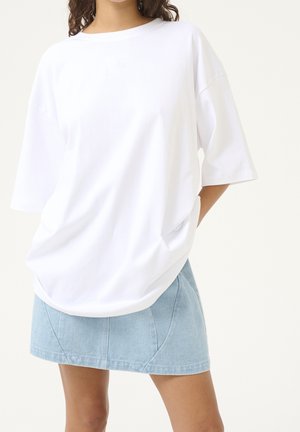 Mujer con una camiseta blanca de manga corta holgada ligeramente metida dentro de una minifalda de mezclilla azul claro, de pie con una mano detrás de la espalda.