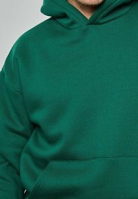 Homme portant un sweat à capuche vert avec une poche avant et une capuche, cadré pour montrer le haut du torse et le bas du visage.