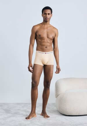 Beżowe bokserki z marka na pasie, model stoi na boso, na jednolitym jasnym tle, pokazując gładką teksturę i dopasowanie.