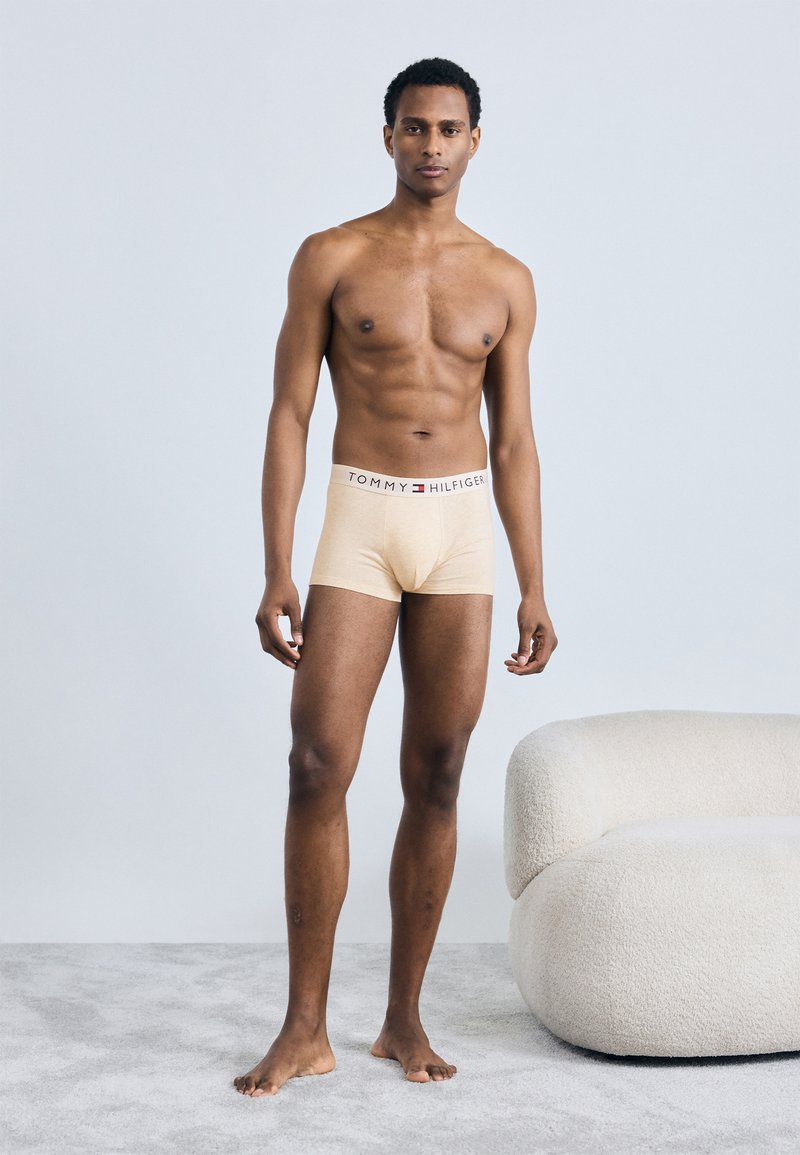 Beżowe bokserki z marka na pasie, model stoi na boso, na jednolitym jasnym tle, pokazując gładką teksturę i dopasowanie.