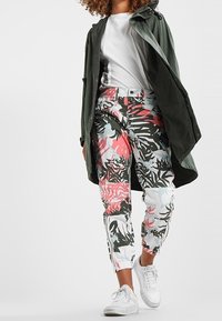 Färgglada, mönstrade joggingbyxor i vitt, med tropiska löv i rosa och grönt, kombinerat med en mörkgrön jacka och vita sneakers.