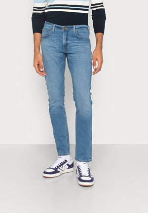 Light blue denim jeans with a slim fit, featuring five pockets and a button closure, paired with white and navy sneakers.