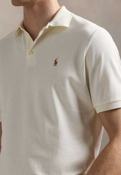 Kortermet poloshirt i lyse kremfarget bomull. Har en klassisk krage, knappestolpe med tre knapper, og en liten brodert logo på brystet.