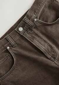 Közeli kép barna farmernadrágról, amely az elülső zsebet, az övhurkokat és a fémgombos rögzítést mutatja.