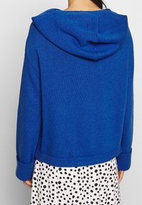 Blå stickad hoodie med avslappnad passform, med en stor huva, ribbade ärmslut och fåll, bärs över en vit klänning med svarta prickar.