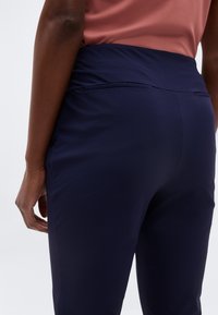 Άτομο που φοράει σκούρο μπλε παντελόνι και ροζ μπλούζα, εμφανιζόμενο από πίσω στο επίπεδο της μέσης και των γοφών, με το χέρι χαλαρό στο πλάι.