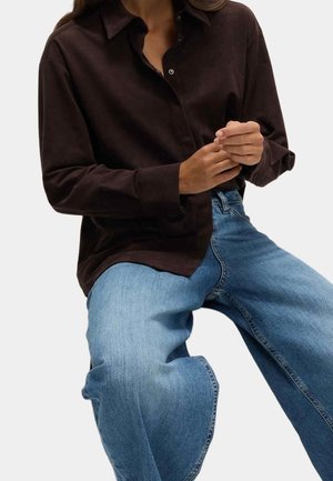 Personne portant une chemise marron foncé boutonnée et un jean bleu clair, assise avec les mains légèrement jointes.