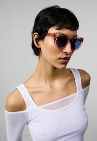 Occhiali da sole oversize di colore rosa con lenti blu, caratterizzati da una forma geometrica a forma di occhio di gatto e una montatura traslucida. Il modello indossa una maglietta bianca.