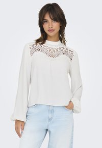 White long-sleeve top with a lace detail around the neckline. Soft fabric, relaxed fit, and gathered cuffs at the sleeves. Worn with light denim jeans.