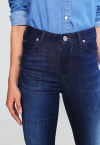 Jeans de mezclilla azul con un diseño ajustado, que presentan una cintura de altura media, cinco bolsillos, cierre de botón de metal y detalles de desvanecimiento sutil.
