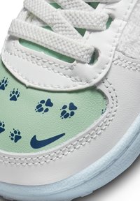 Bílé tenisky s mátově zelenými detaily, s modrými vzory tlapek, texturovanými materiály a bílými tkaničkami. Obsahuje logo Nike swoosh.