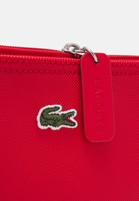 Sacchetto di tessuto rosso con una superficie texturizzata, una cerniera argentata e una toppa con logo di coccodrillo verde, con un'etichetta rossa Lacoste attaccata.