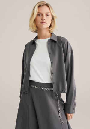 Gray button-up shirt with a relaxed fit and long sleeves, worn over a white t-shirt. Features a chain belt detail on gray trousers.