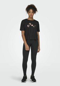 La camiseta negra corta presenta un patrón abstracto en rosa, combinada con leggings negros y zapatillas deportivas negras. Tela suave, corte relajado.
